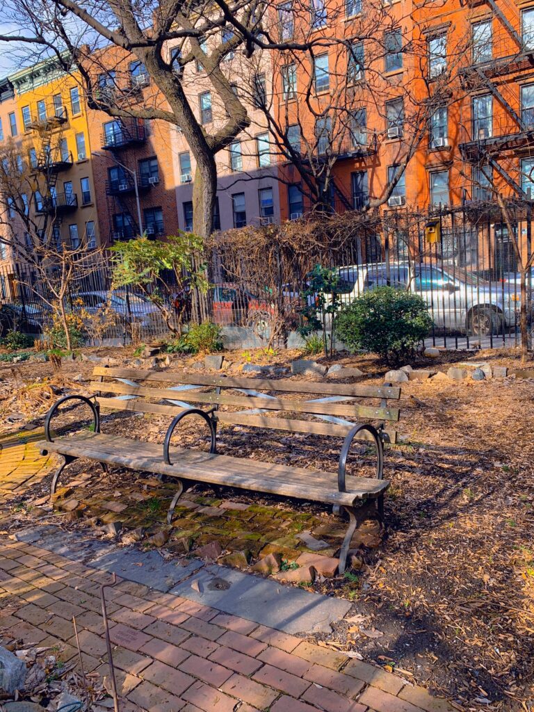 Wooden bench on dirt covered ground. In the back there is an iron fence and side view of colorful apartment buildings. In front of the bench is a brick and cement pathway. 